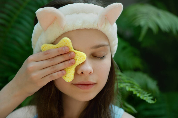 Beautiful little kid with funny bunny headband cleansing her face by yellow star  shape sponge. Portrait of cute little teenage girl enjoying skin care procedures. - Foto, imagen
