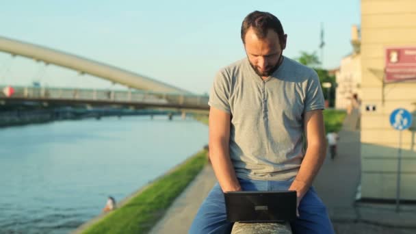 Man with laptop sitting on wall by river - Záběry, video