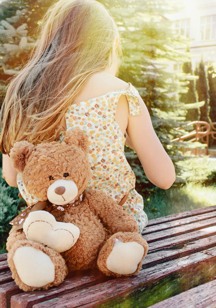 little girl sitting back with teddy bear on the bench in sun bea - Photo, image
