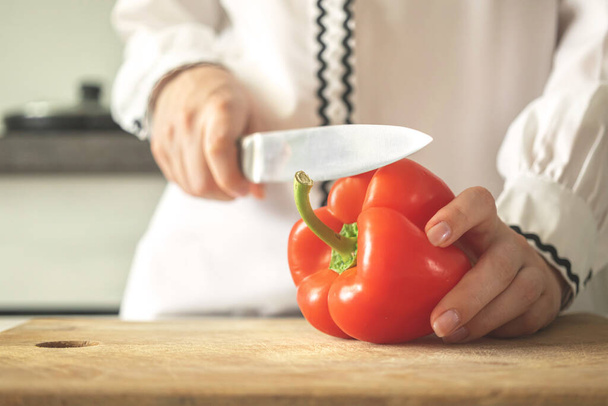 Chef slicing sweet pepper on wooden board, modern kitchen background. Young woman preparing sweet bell pepper for healthy food - 写真・画像