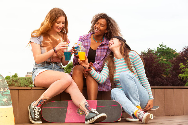 Three teenager skater friends, afro and caucasian, skating and drinking smoothies in a park - Photo, Image