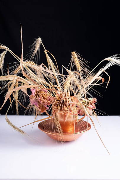 a bouquet of wheat ears with dry flowers in a copper dish on a white table and black background - Fotografie, Obrázek