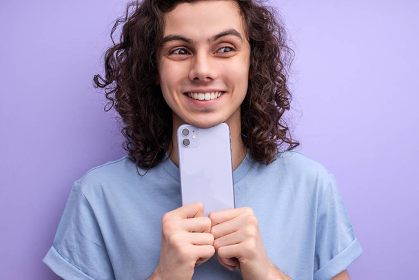 Молодой улыбающийся парень фотографирует на смартфоне, глядя в сторону - Фото, изображение