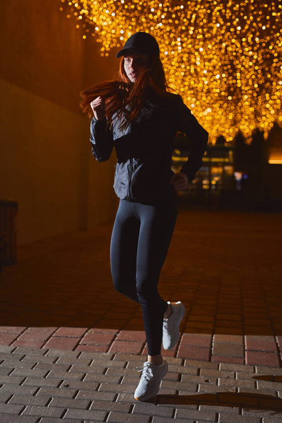 athlete redhead woman in sportswear running along road in city at night, sport concept - Фото, изображение