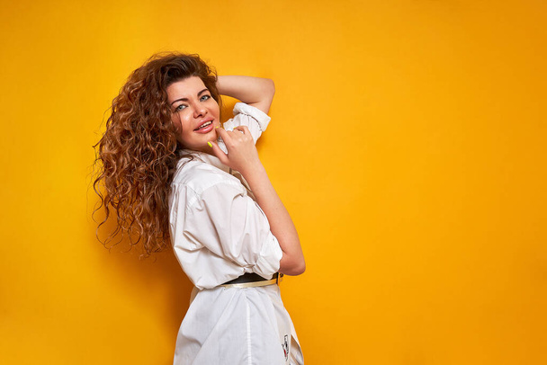 young curly haired woman in white shirt holds her hand to chin and adjusts her hair with thoughtful face stands sideways isolated on yellow background with place for text thinking about right decision - Photo, Image