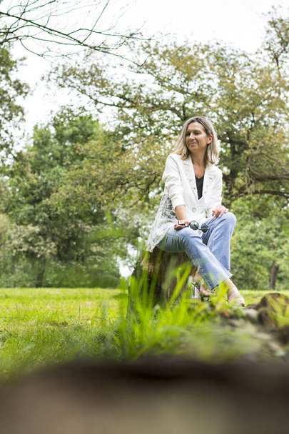 Mujer posando sentada sobre un tronco de madera en un parque de Asturias, España.La niña lleva vaqueros, chaqueta blanca y zapatos transparentes.Foto con un desenfoque en primer plano y rodada en formato retrato. - Foto, Imagen