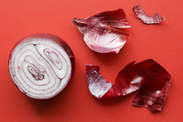 Una mitad cortada de una cebolla roja yace sobre un fondo rojo junto a una cáscara de cebolla pelada. Primer plano - Foto, Imagen