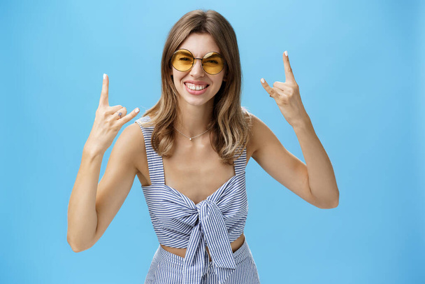 Tyttö valmis rock-n-roll tämä osapuoli yllään paras matching asu vaatekaappi ja viileä aurinkolasit osoittaa rock eleitä, hymyillen innoissaan hauskaa kuunnella musiikkia konsertissa yli sininen seinä - Valokuva, kuva