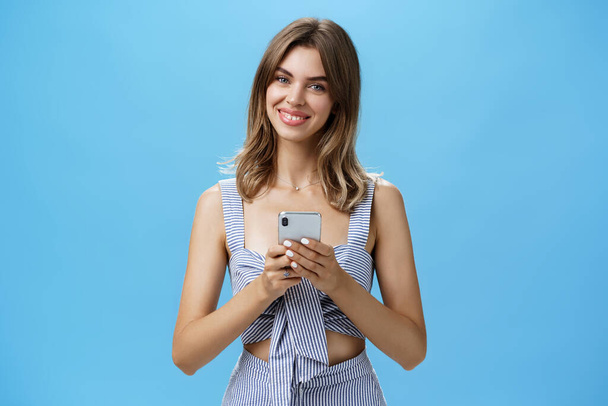 Přátelský příjemný dobře vypadající žena v odpovídající oblečení drží smartphone přes hrudník naklápění hlavy s úsměvem široce ukazující roztomilé mezery zuby jsou potěšeni po přečtení srdcervoucí zprávu - Fotografie, Obrázek