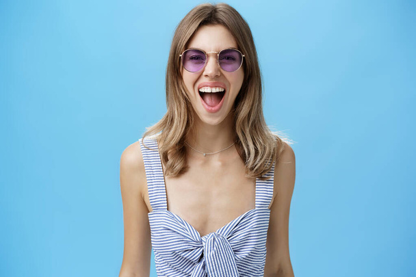 Expressive and careree woman being wild ad free yelling out loud at camera wearing awesome stylish sunglasses and striped top letting out emotions being open-minded and rebellious over blue wall - Fotoğraf, Görsel