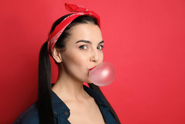 Fashionable young woman in pin up outfit blowing bubblegum on red background, space for text - Фото, изображение