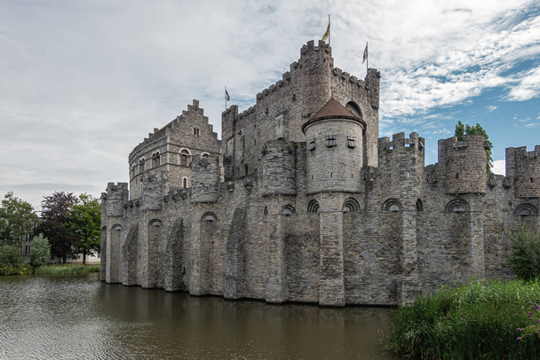 Gent, Flanders, Belgium - July 30, 2021: West side of gray Gravensteem medieval stone castle under blue cloudscape behind dark water moat. Some green foliage. - Photo, Image