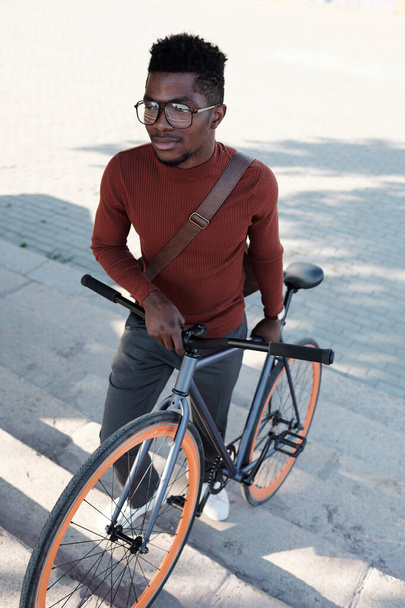Νεαρός επιχειρηματίας με γυαλιά ηλίου και έξυπνο καθημερινό ντύσιμο που σπρώχνει ποδήλατο - Φωτογραφία, εικόνα