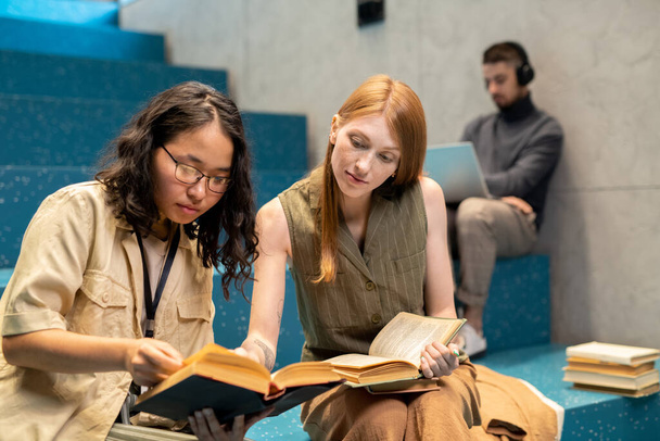 Δύο νεαρές γυναίκες με καθημερινά ρούχα συζητούν για βιβλία εναντίον ενός τύπου με φορητό υπολογιστή - Φωτογραφία, εικόνα