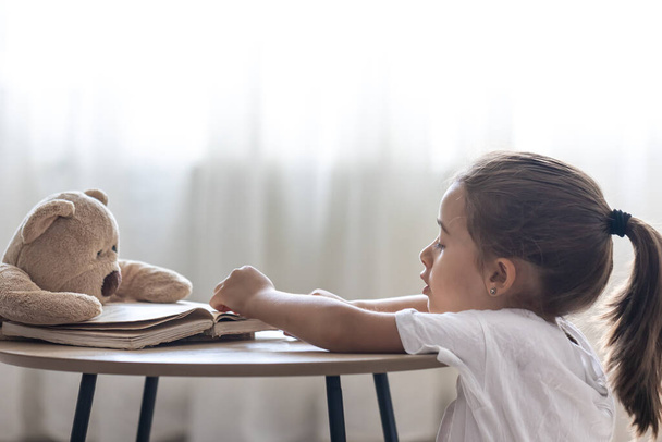 Ένα κοριτσάκι παίζει με το αρκουδάκι της και ένα βιβλίο, του μαθαίνει να διαβάζει, παίζει στο σχολείο.. - Φωτογραφία, εικόνα