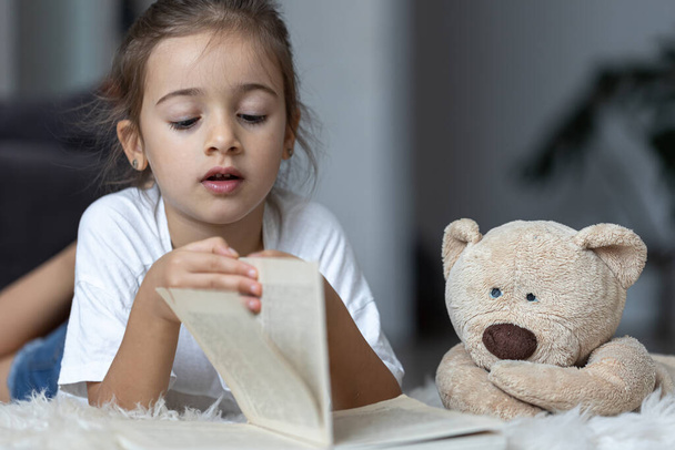 Cute little girl w domu, leżąc na podłodze z jej ulubioną zabawką i czyta książkę. - Zdjęcie, obraz