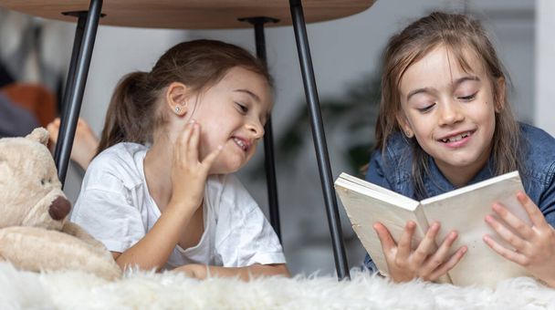 Dos hermanas pequeñas se divierten leyendo un libro juntas mientras yacen en el suelo en su habitación. - Foto, imagen