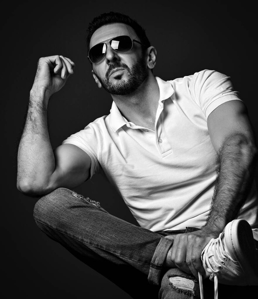 Black and white. Stylish adult unshaved man in white polo shirt, jeans and modern sunglasses sits with legs crossed - Foto, Bild