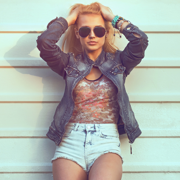 Outdoor summer closeup portrait of young stylish fashion glamorous woman or girl posed in  sunny day on street jeans jacket and sunglasses standing near white wall - Photo, Image