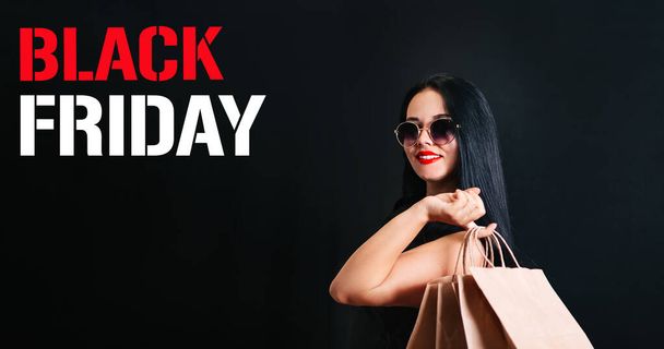 un hermoso cliente con una bolsa de compras en sus manos y gafas oscuras, aislado sobre un fondo negro en las vacaciones del Viernes Negro. Espacio para texto - Foto, Imagen