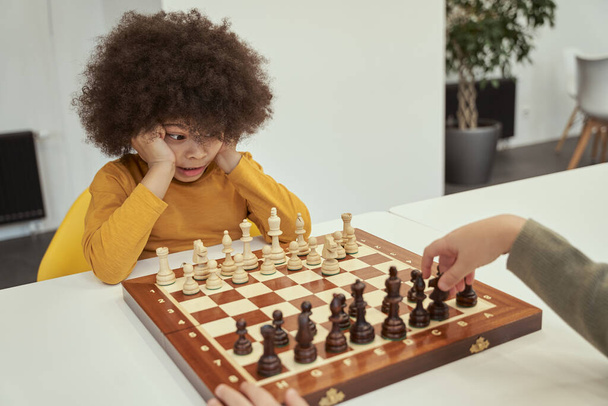 Bel giocatore. Divertente ragazzino con i capelli afro guardando il suo amico fare una mossa mentre gioca a scacchi, seduto al tavolo al coperto - Foto, immagini