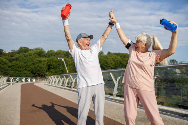 Cheerful senior man and woman raise up bottles and join hands on footbridge - Photo, Image