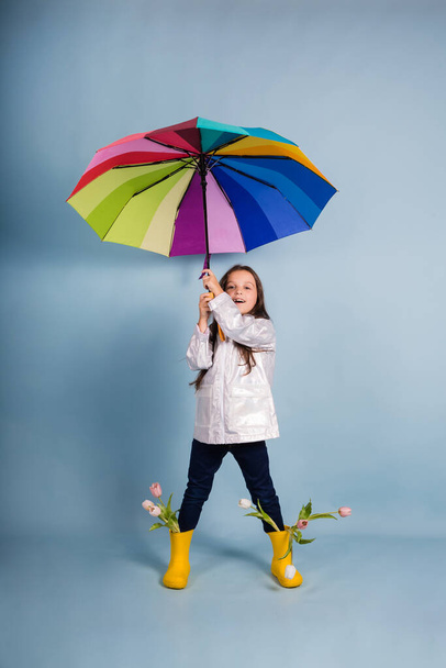 a funny little girl in a raincoat and yellow rubber boots holds a multi-colored umbrella on a blue background - Foto, Imagen