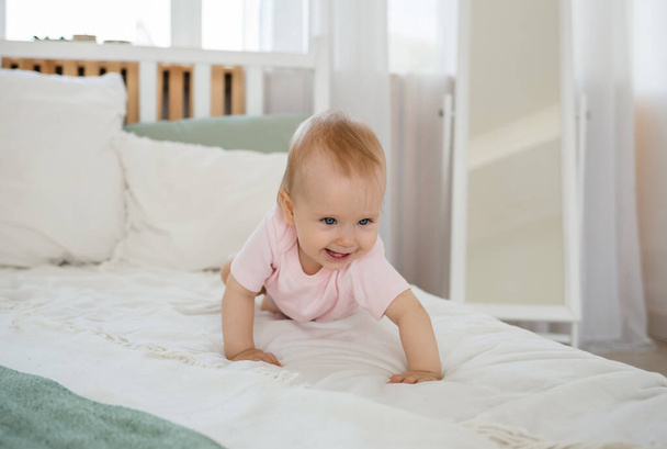 baby meisje in een roze bodysuit kruipt op het bed in de kamer - Foto, afbeelding