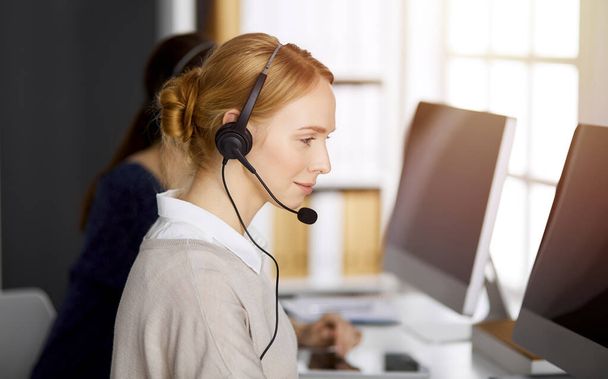 Friendly caucasian businesswoman talking by headset in sunny office. Call center and diverse people group in business - Fotó, kép