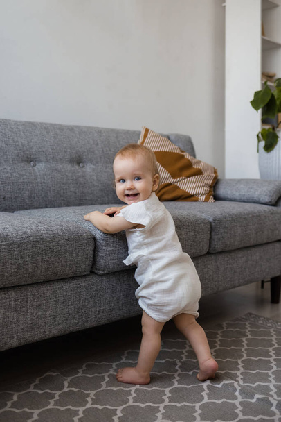 una bambina in tuta mussulmana è in piedi accanto a un divano grigio nella stanza e sorridente - Foto, immagini