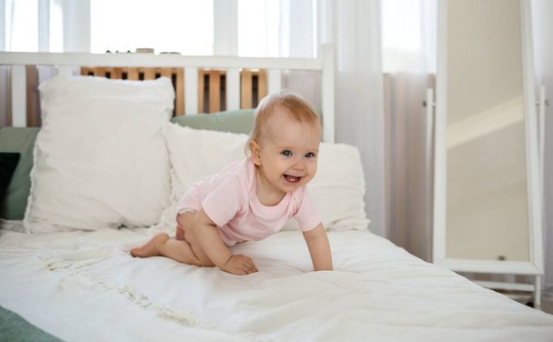 Pembe elbiseli küçük kız pamuklu çarşafların üzerinde sürünüyor odadaki yatakta. - Fotoğraf, Görsel