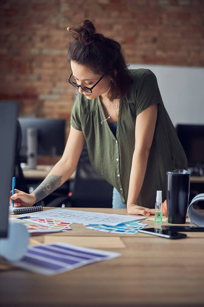 Σχεδιαστής εσωτερικών χώρων ή αρχιτέκτονας σε casual φόρεμα με βρώμικο χτένισμα κρατώντας σημειώσεις, ενώ εργάζονται σε νέο έργο, στέκεται στο γραφείο της την ημέρα - Φωτογραφία, εικόνα