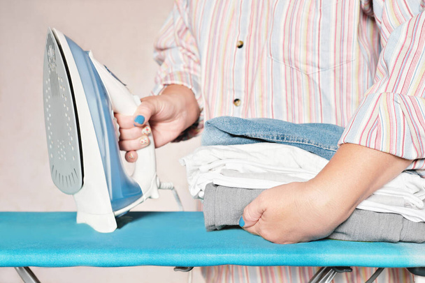 pila de ropa ropa y mujer sosteniendo hierro. trabajo en casa, tareas, concepto de limpieza. blanco gris azul textiles ropa - Foto, Imagen