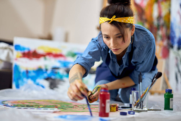 Jeune femme concentrée, peintre dans le tablier choisir la couleur de la peinture, tenant le pinceau tout en travaillant sur la peinture, assis sur le sol à la maison atelier - Photo, image