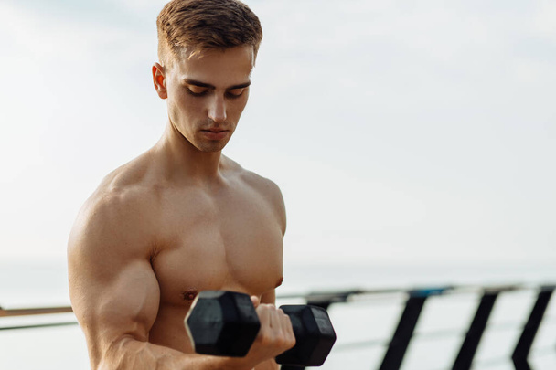 Muscular handsome strong man with naked torso exercising with dumbbells showing six abs cubes outdoors on the bridge on the pier - Photo, Image