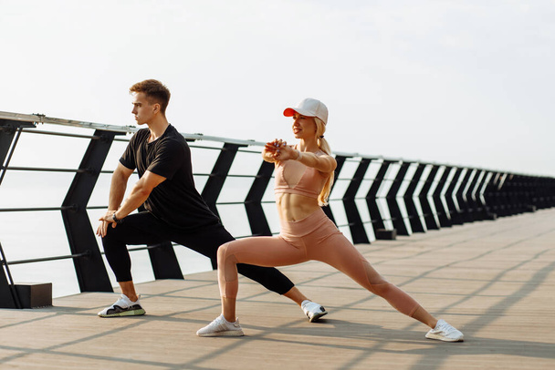 Attractive young fitness couple doing stretching exercises outdoors on the sea on the bridge, sportive man and woman exercising together - Fotó, kép