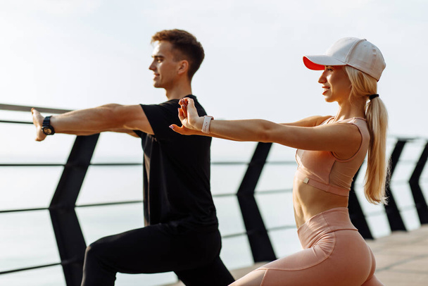 Attractive young fitness couple doing stretching exercises outdoors on the sea on the bridge, sportive man and woman exercising together - Photo, image