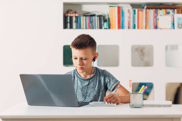 Jonge jongen in headset zit aan tafel met laptop en bereidt zich voor op school. Online onderwijsconcept. - Foto, afbeelding
