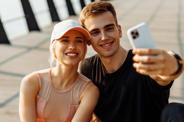Atraktivní mladí sportovci muž a žena odpočívající po cvičení s selfie na mobilním telefonu sedí na fitness podložce venku na molu - Fotografie, Obrázek