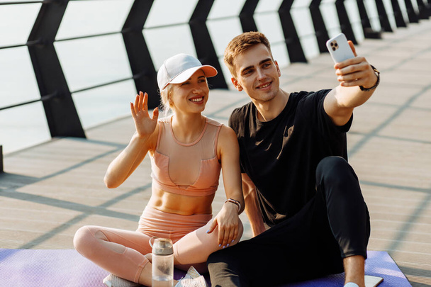 Attractive young sportsmen man and woman resting after workout taking selfie on mobile phone sitting on fitness mat outdoors on pier - Valokuva, kuva