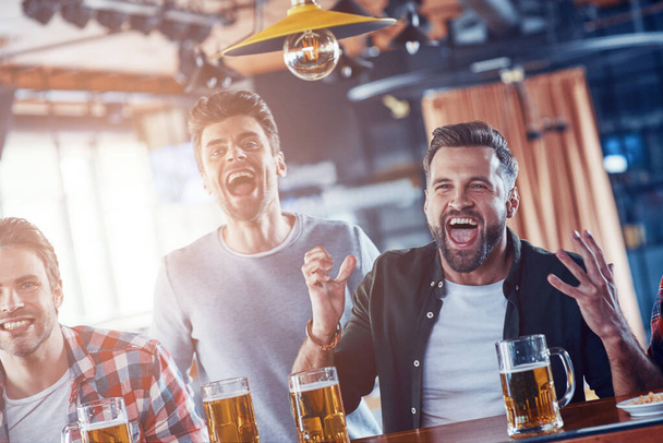 Cheering young men in casual clothing drinking beer and watching sport game while sitting in the pub - Foto, Imagem