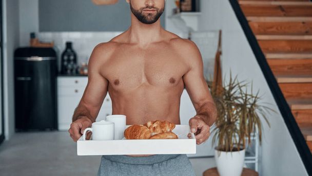 Good looking young shirtless man holding tray with breakfast while spending time at home - Fotografie, Obrázek