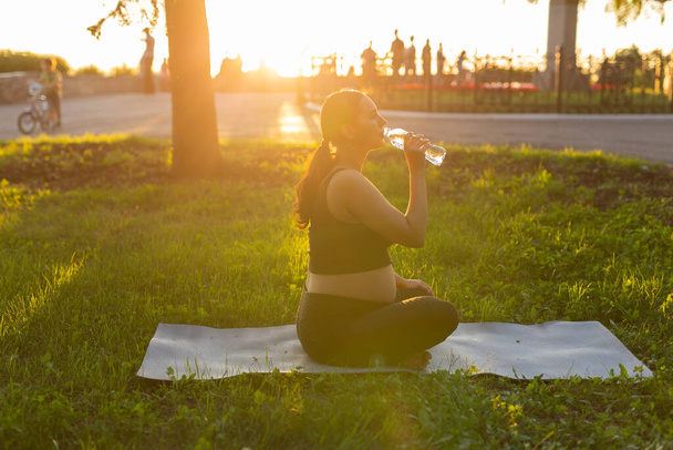 Grossesse, sport et mode de vie sain - femme enceinte buvant de l'eau à partir d'une bouteille assise sur l'herbe au soleil jour d'été après le yoga - Photo, image