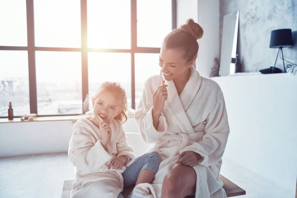 Мати і дочка в халатах посміхаються і чистять зуби під час ранкової рутини
 - Фото, зображення