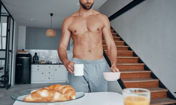 Good looking young shirtless man having breakfast while spending time at home kitchen - 写真・画像