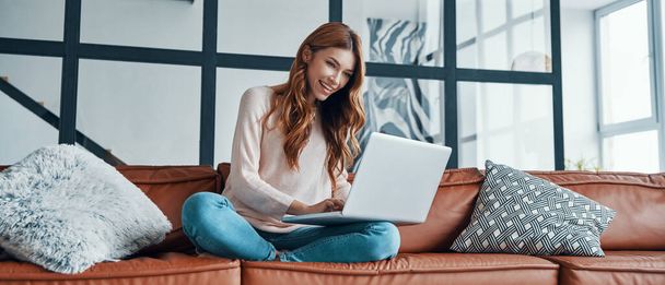 Attractive young woman using laptop and smiling while spending time at home - Zdjęcie, obraz