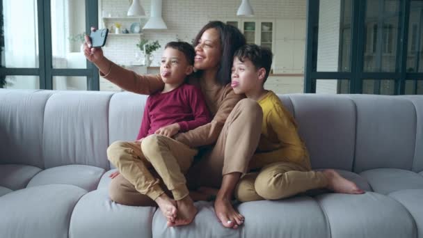 Grappige vrolijke Afro-Amerikaanse vrouw maakt selfie ptotos, houdt een mobiele telefoon in uitgestrekte armen, knuffelt zachtjes haar schattige zonen en grimmig kijkend naar de camera. - Video