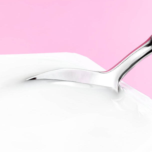 Copa de yogur y cuchara de plata sobre fondo rosa, recipiente de plástico blanco con crema de yogur, productos lácteos frescos para una dieta saludable y equilibrio nutricional - Foto, Imagen