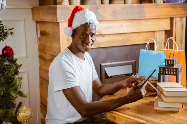 afro americano bonito cara no Santa chapéus estão usando smartphones faz férias saudações e sorrindo, homem comprar coisas on-line shoping com seu telefone - Foto, Imagem
