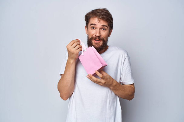 Χαρούμενος άνδρας σε λευκό t-shirt με ροζ μικρό πακέτο δώρου φως φόντο - Φωτογραφία, εικόνα